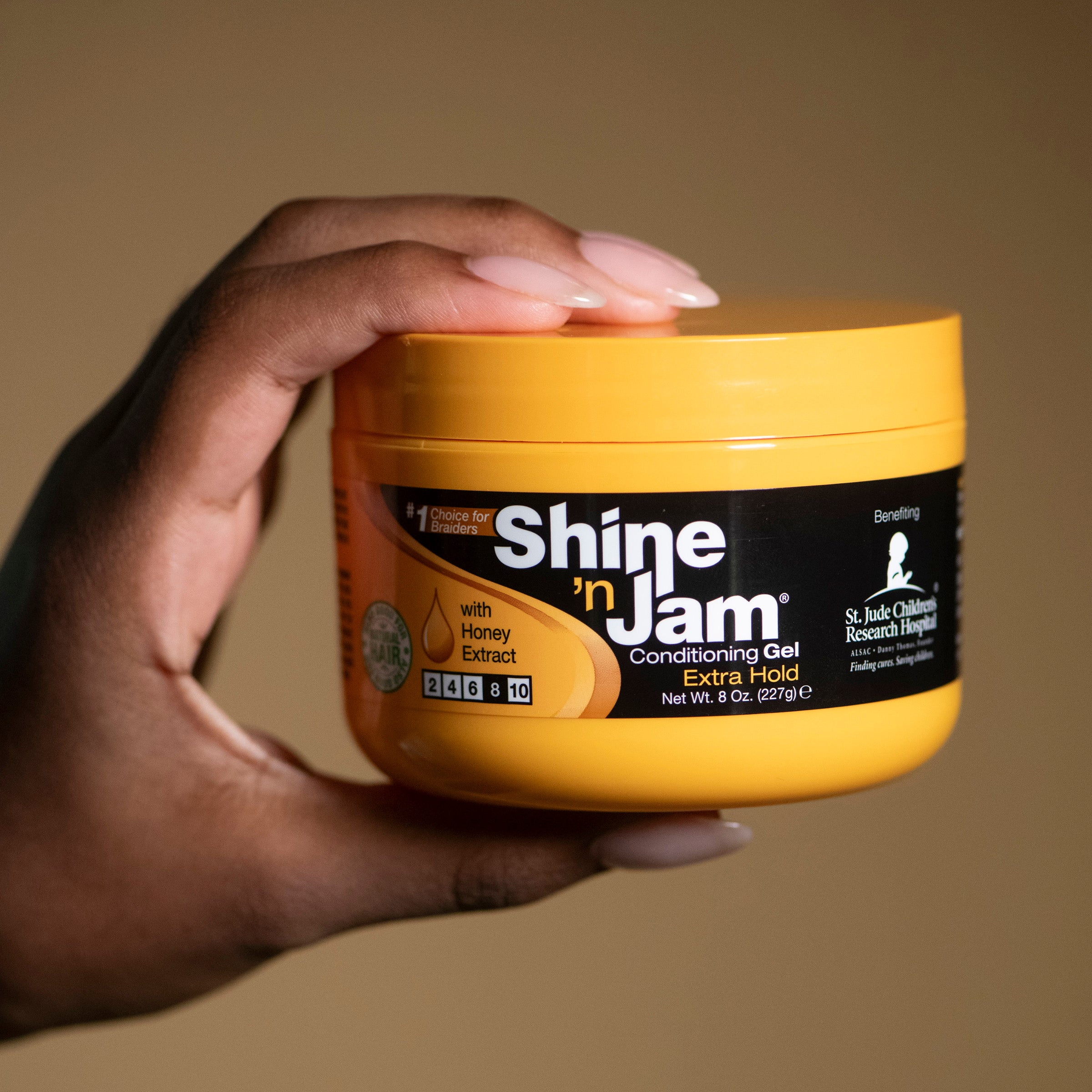 En hånd holder en gul krukke med Ampro Shine 'n Jam Conditioning Gel. Krukken har sort skrift og et billede af en honningkam. Produktet er designet til at give håret ekstra hold og glans.