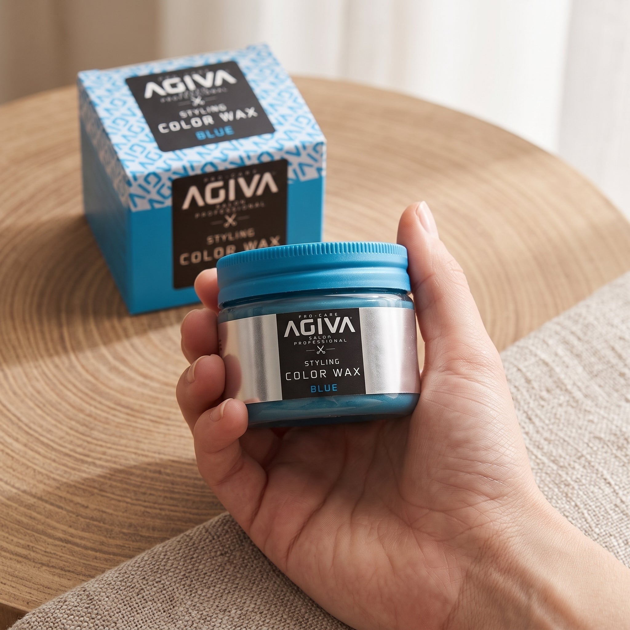 Hånd holder AGIVA Styling Color Wax i blå farve, med produktæske i baggrunden på træbord.