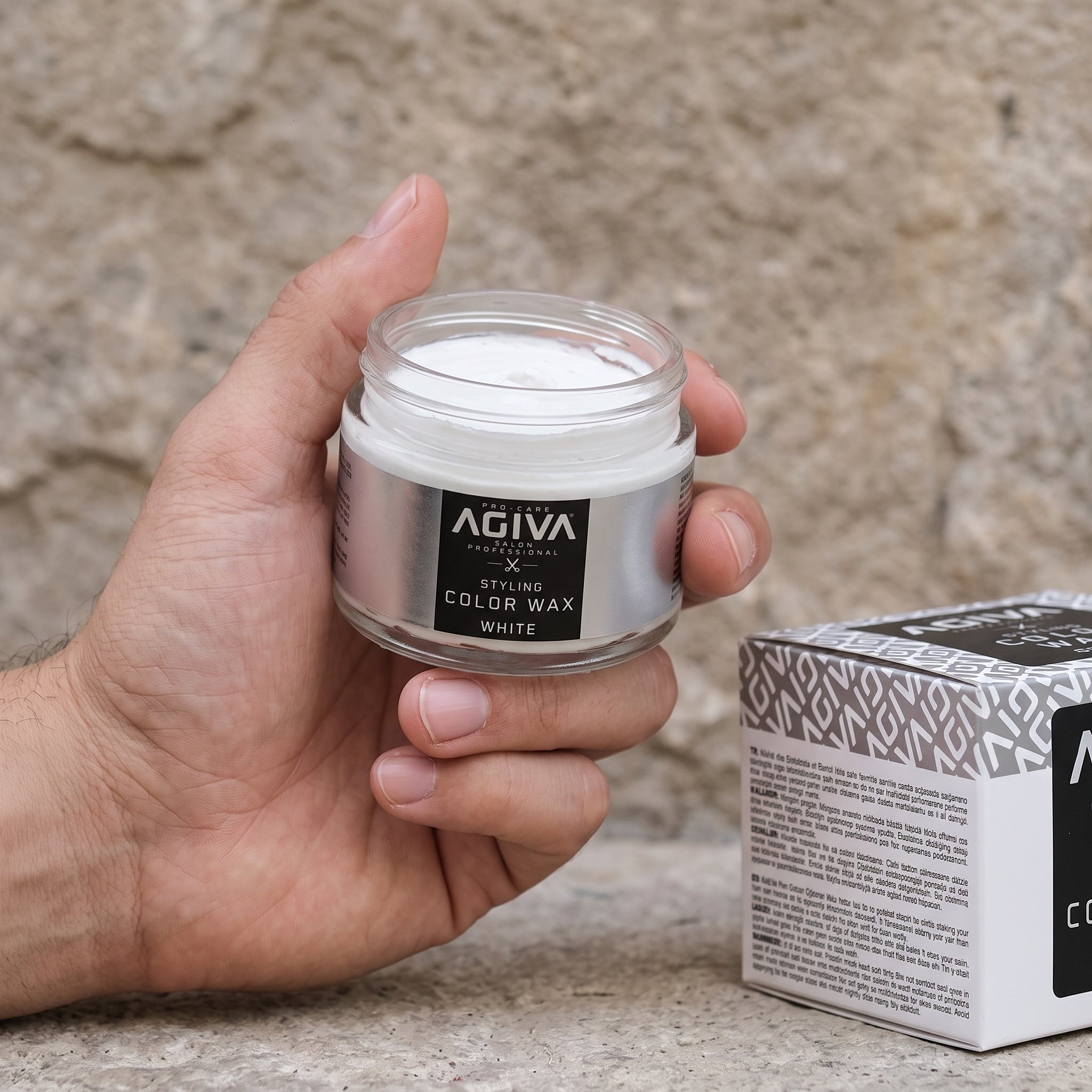 Hånd holder åben glaskrukke med Agiva Styling Color Wax White ved siden af æske på stenunderlag.