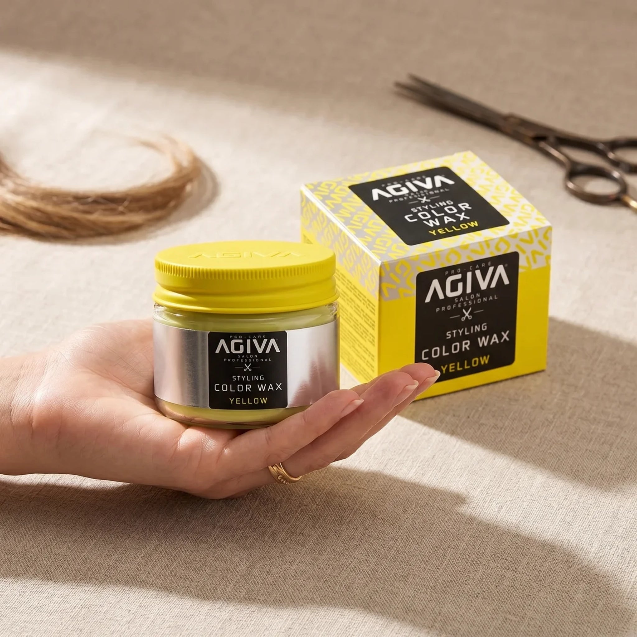 Hånd holder AGIVA Styling Color Wax Yellow i glas med gul låg ved siden af æske, saks og hårlok.