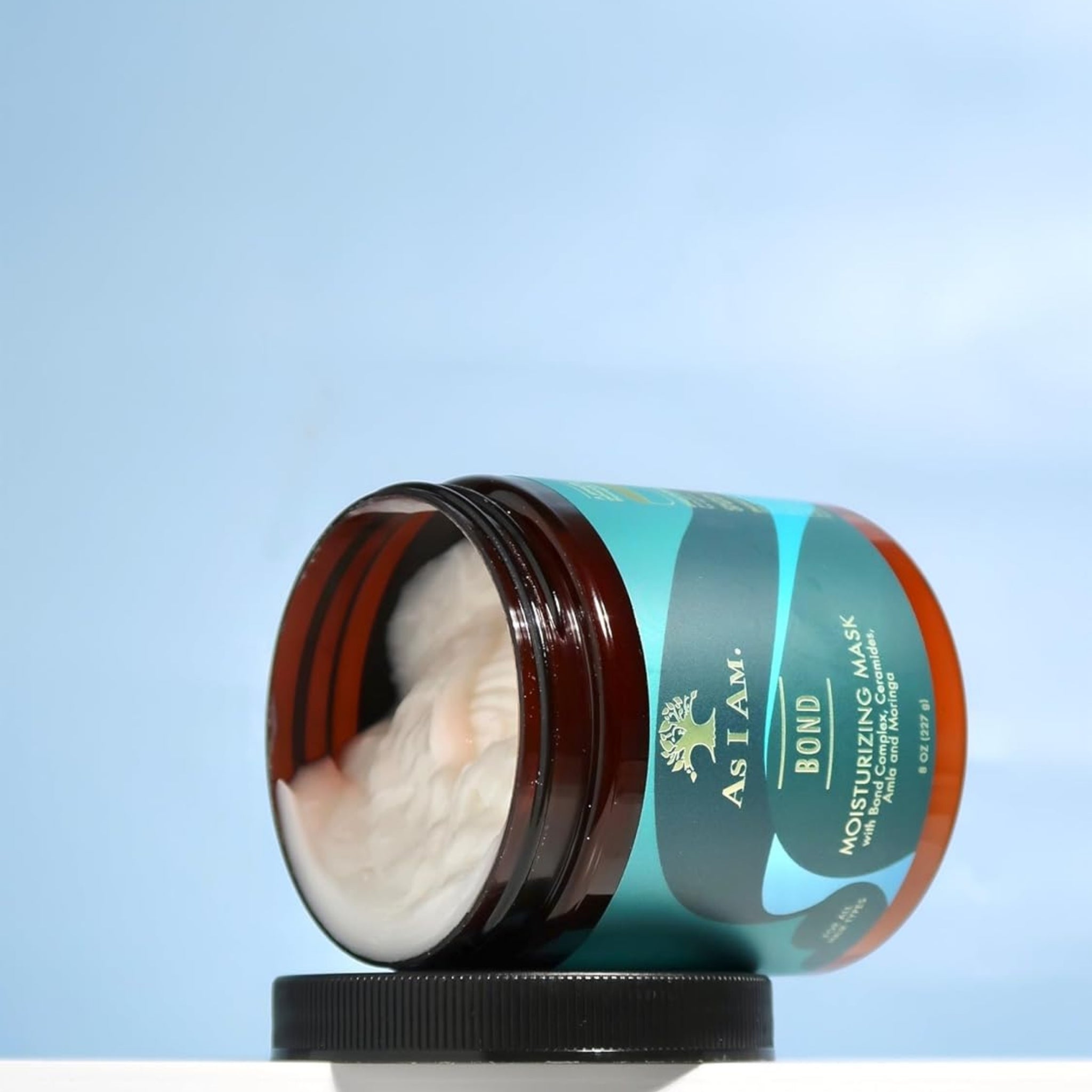As I Am Bond Moisturizing Mask i åben brun krukke med cremet hvid hårmaske, lyseblå baggrund.