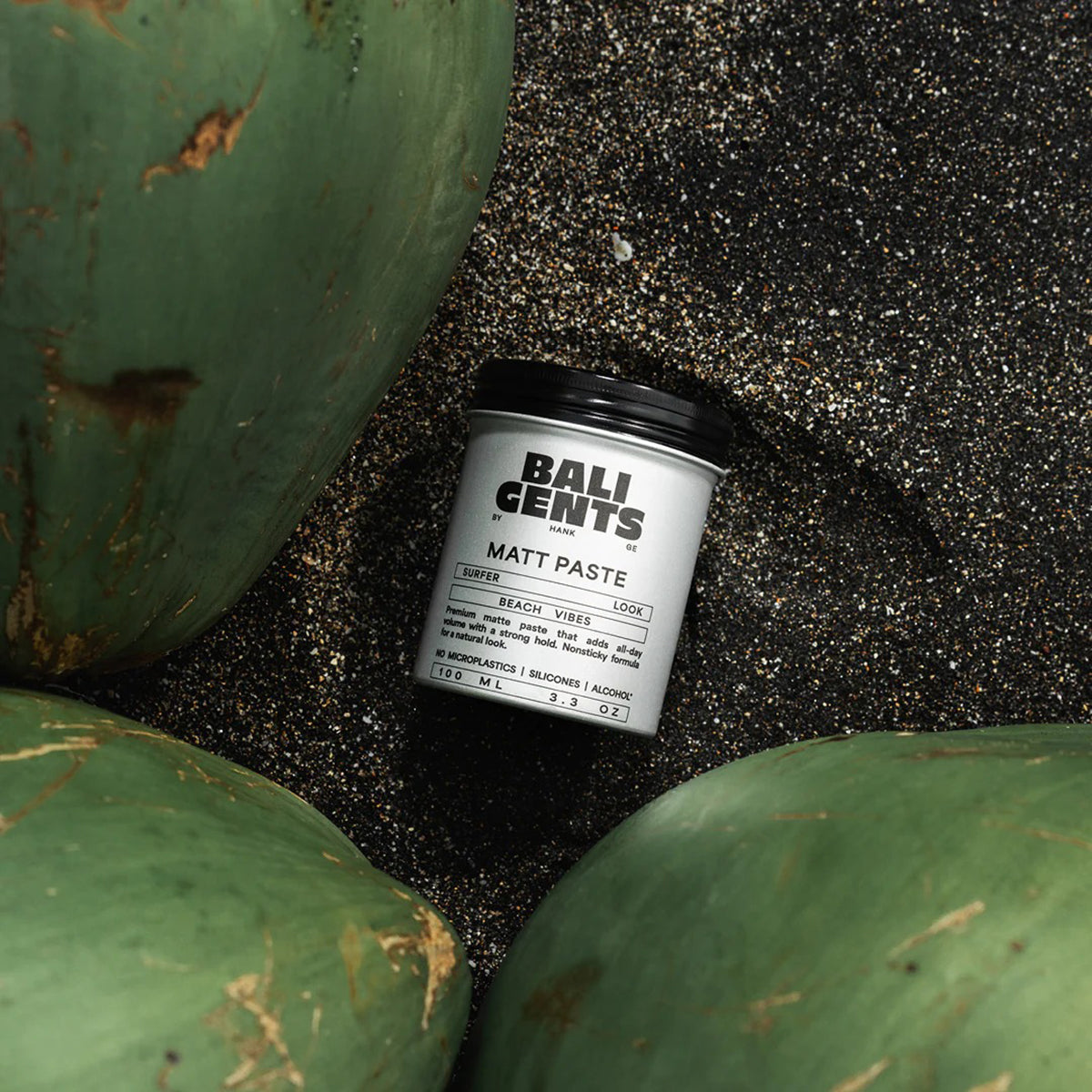 Bali Gents Matt Paste 100ml placeret på sort sandstrand omgivet af grønne kokosnødder giver en naturlig beach vibe.