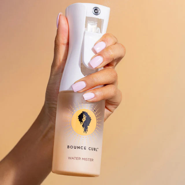Hånd holder Bounce Curl Water Mister med hvid spraydyse og logo foran beige baggrund.