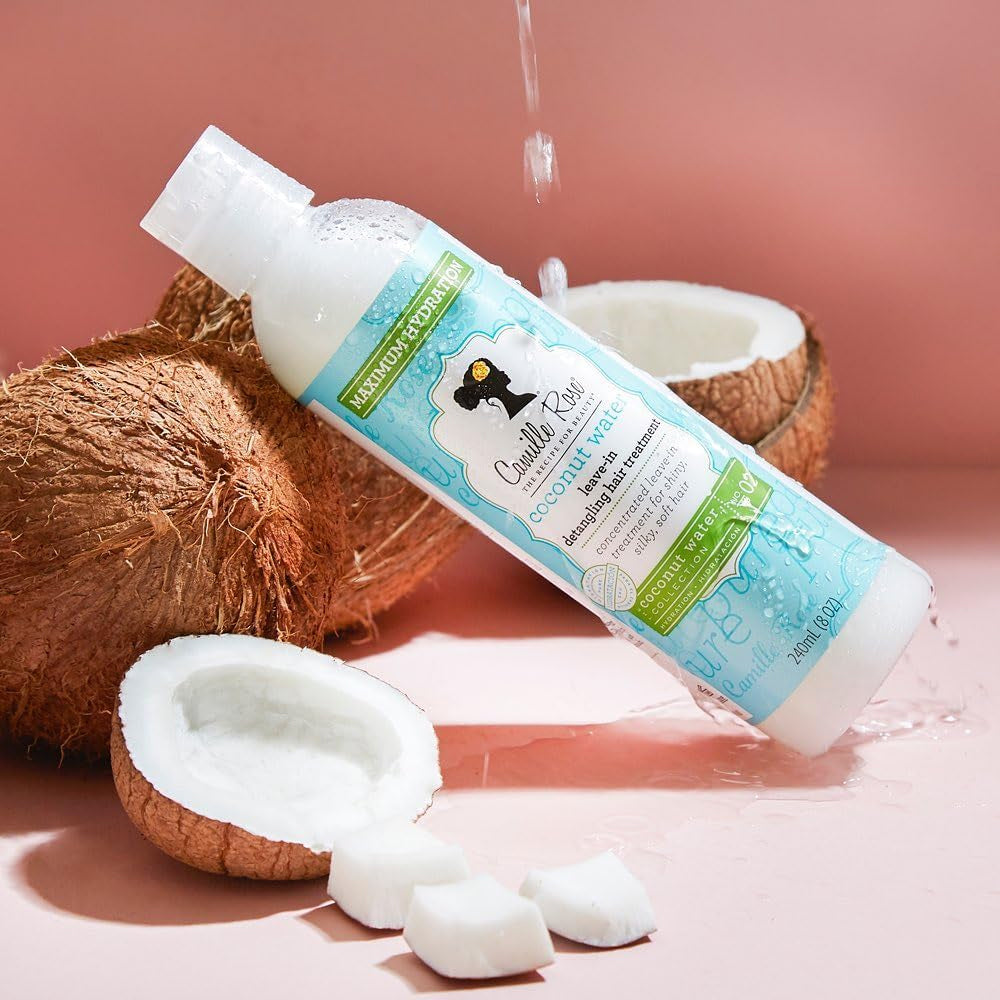 Hvid flaske med Camille Rose Coconut Water Leave-In Treatment ligger ved siden af en åben kokosnød med vanddråber.
