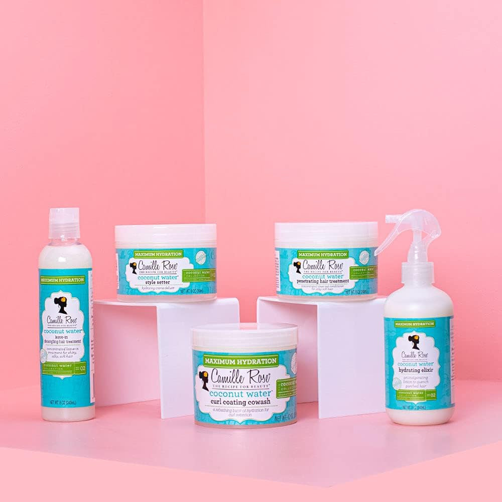 Camille Rose Coconut Water Penetrating Hair Treatment, Curl Coating Cowash og andre produkter på hvide podier mod lyserød baggrund.