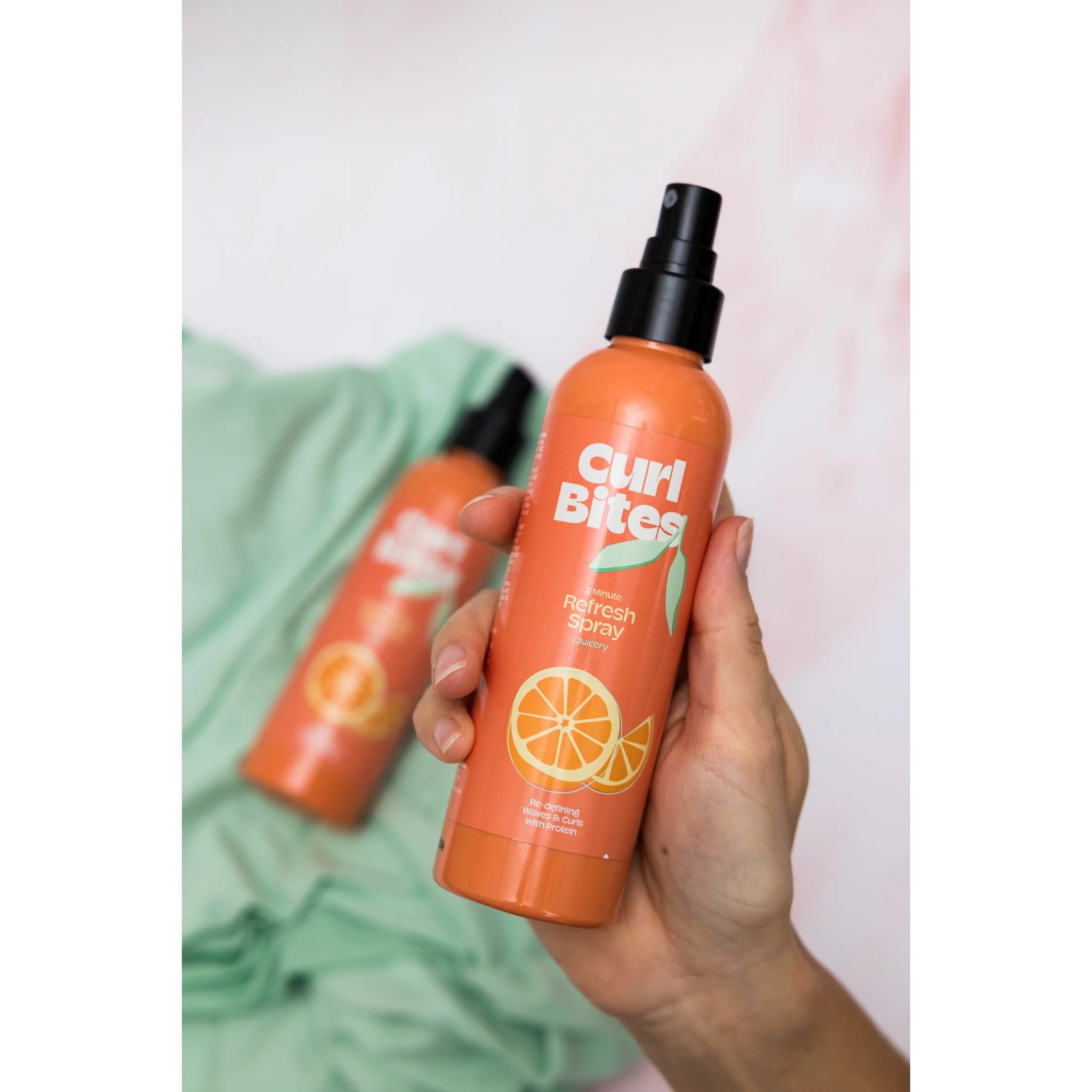 Hånd holder Curl Bites 2 Minute Refresh Spray, orange flaske med appelsinmotiv, mod grøn baggrund.