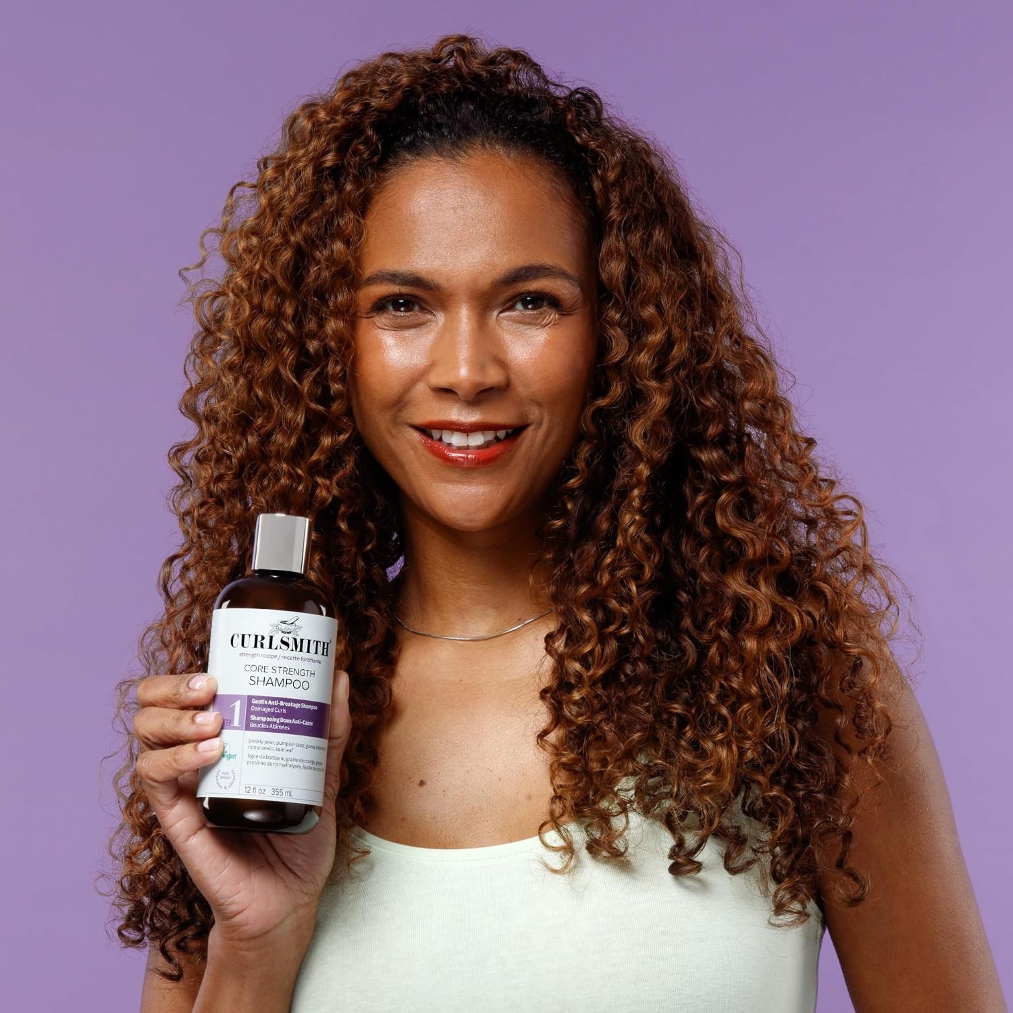 Kvinde med krøllet hår smiler og holder Curlsmith Core Strength Shampoo mod lilla baggrund.