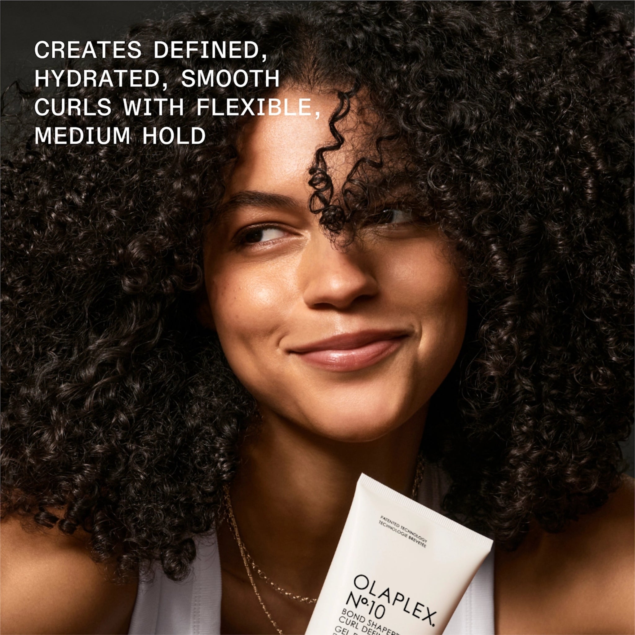 Smilende kvinde med definerede krøller holder Olaplex No.10 Bond Shaper Curl Defining Gel til hydrerede krøller med fleksibel hold.