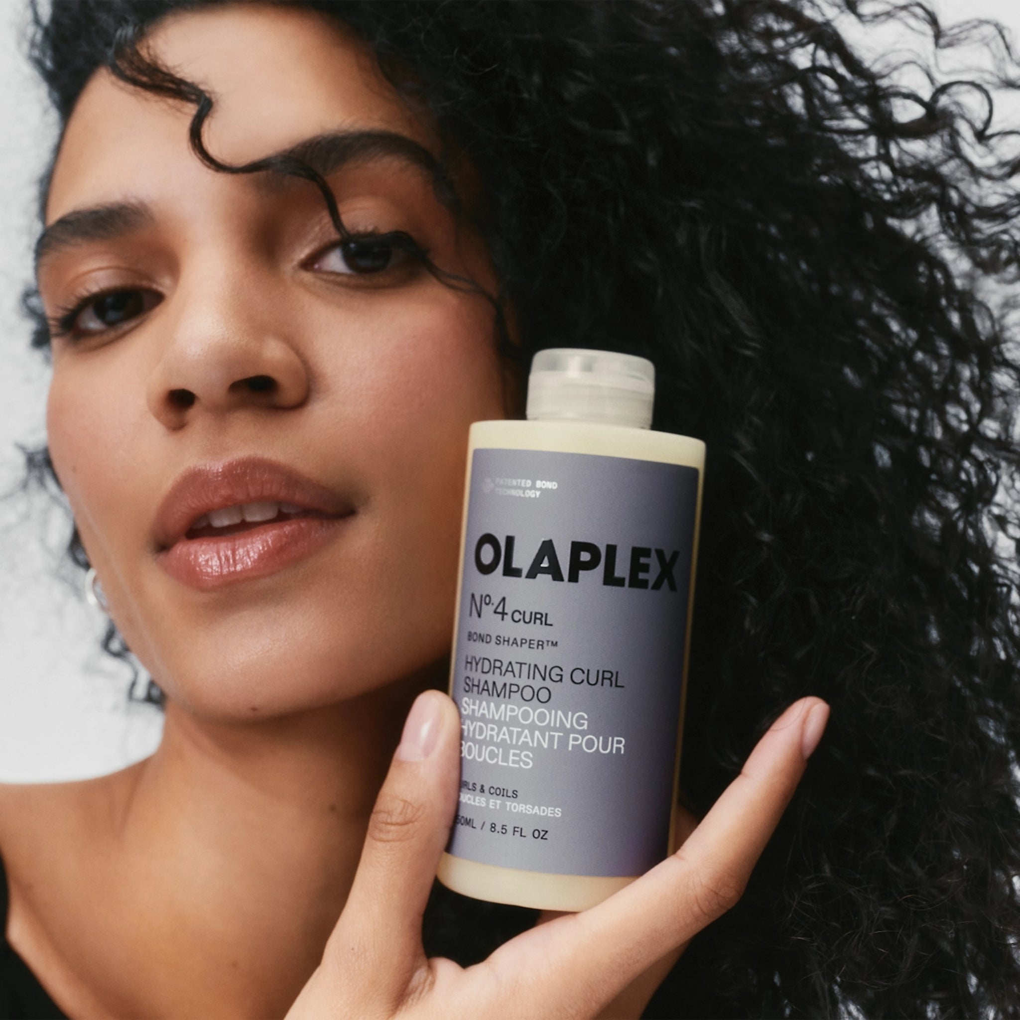 Kvinde med mørke krøller holder Olaplex Nº.4 Curl Bond Shaper hydrating curl shampoo tæt på ansigtet.