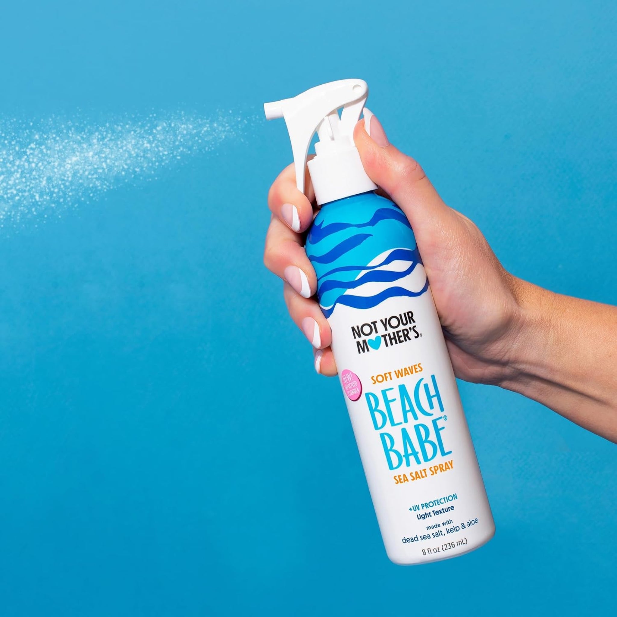 Hånd der sprøjter Not Your Mother's Beach Babe sea salt spray mod blå baggrund.
