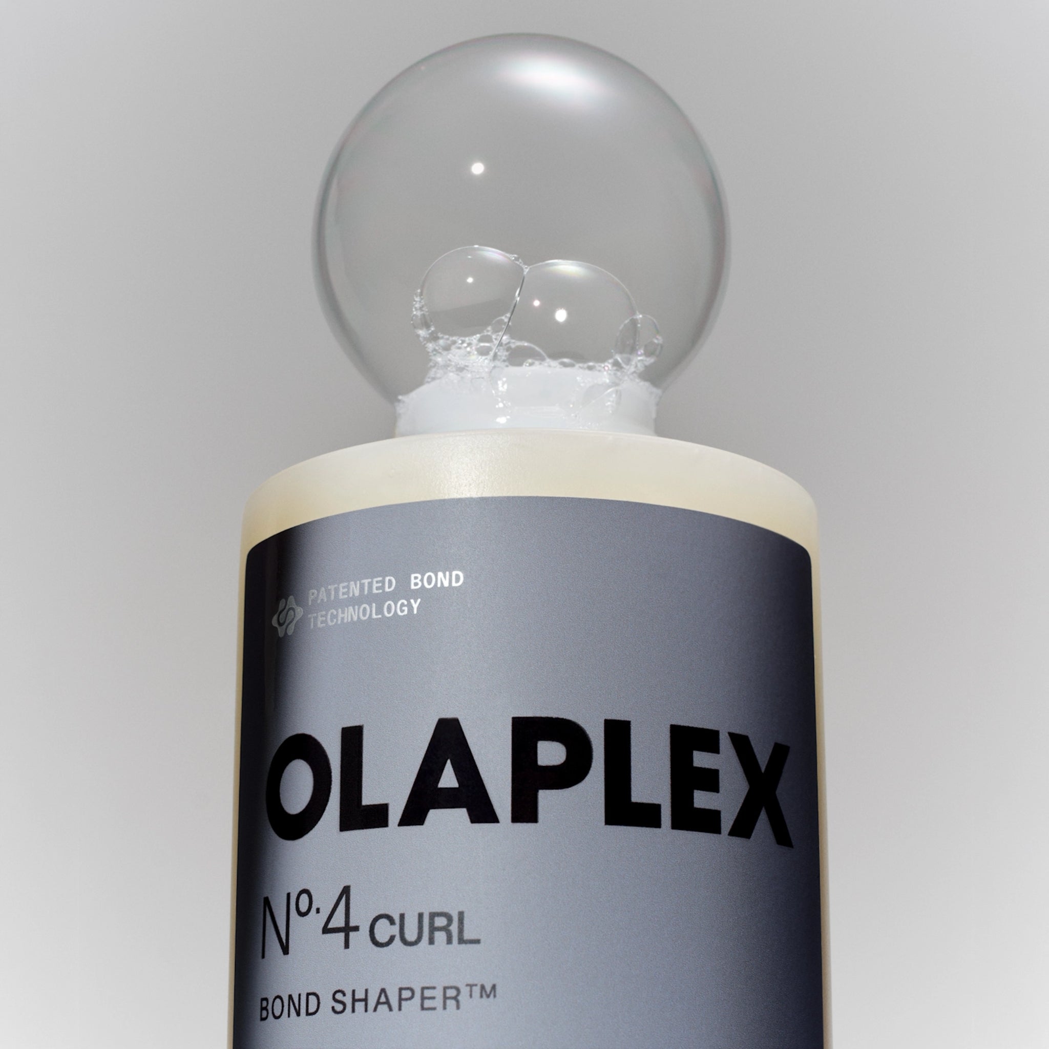 Olaplex No.4 Curl Bond Shaper med gennemsigtig kuglelåg og skum på flaskens top mod grå baggrund.