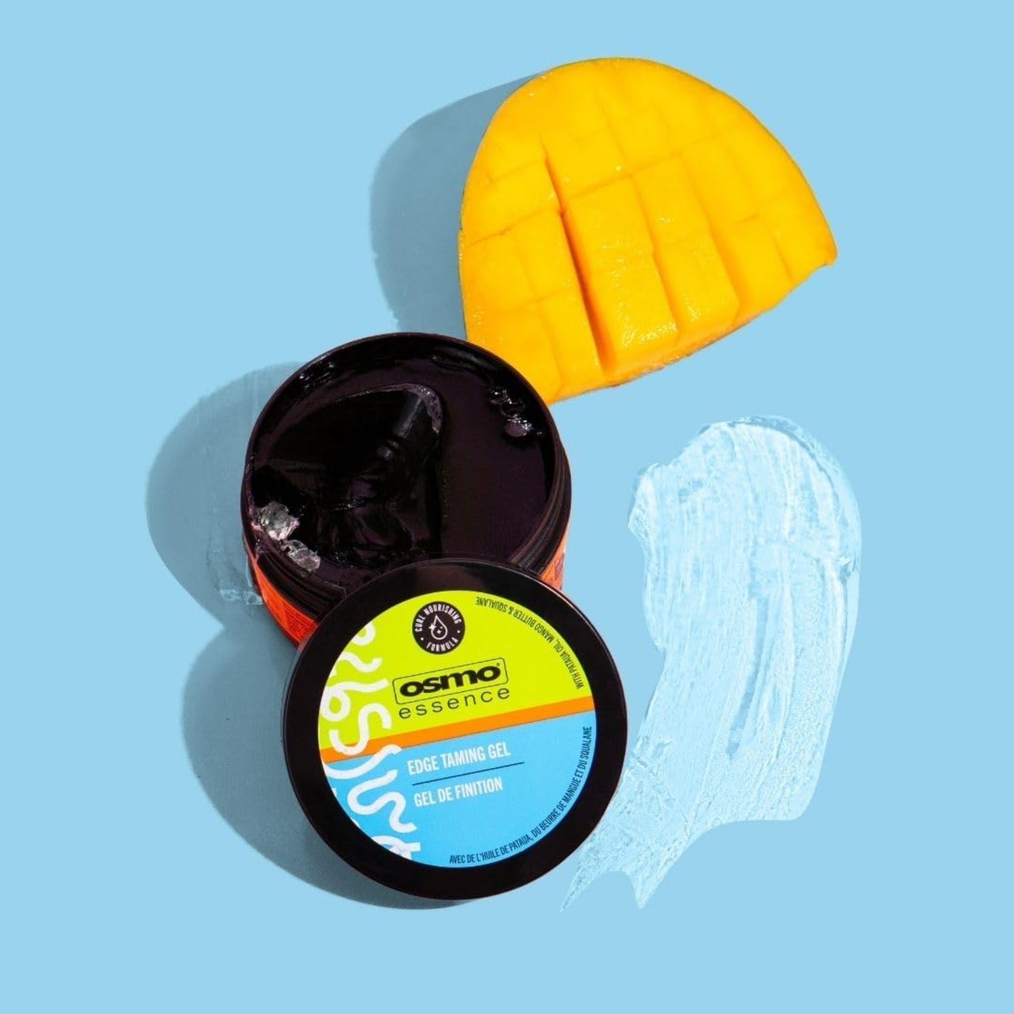 Osmo Essence Edge Taming Gel i åben sort dåse ved siden af udskåret mango på lyseblå baggrund.
