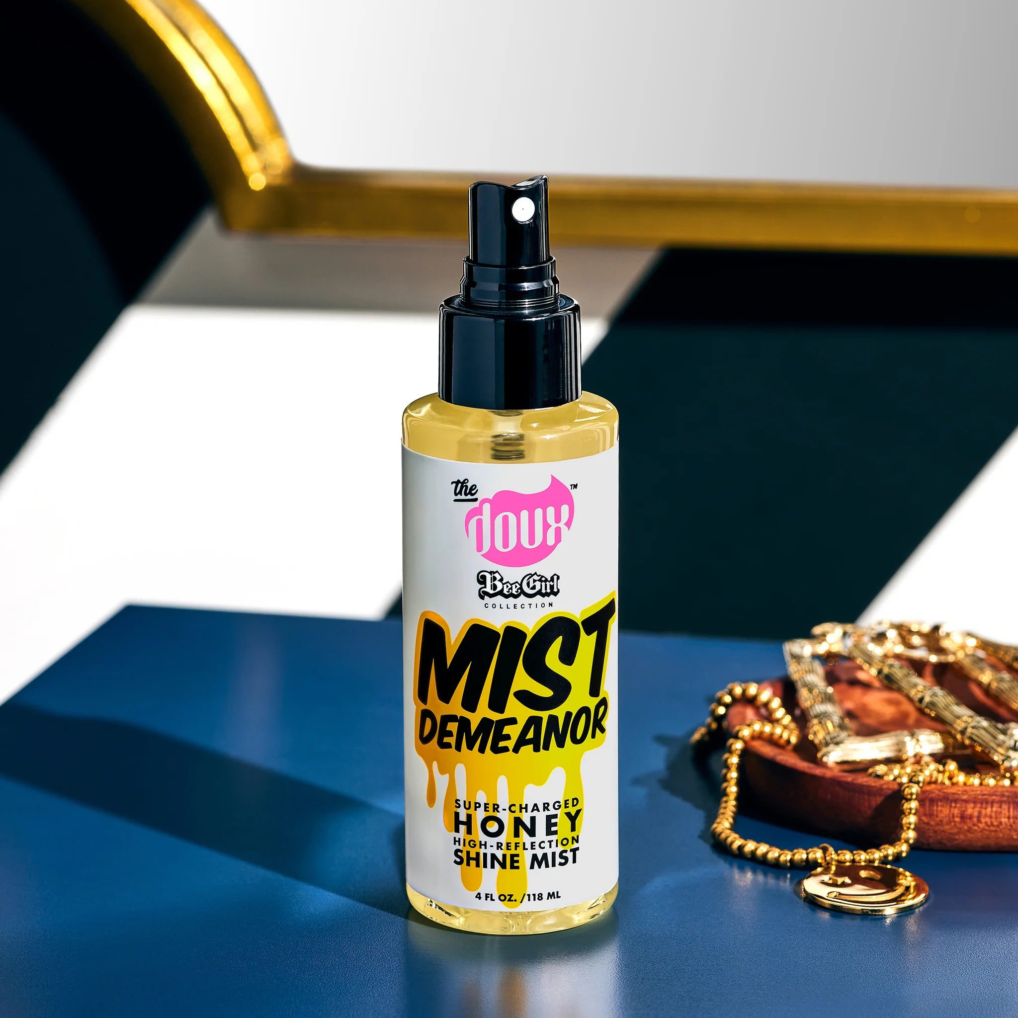 The Doux Mist Demeanor honey shine mist-flaske med sort spraypumpe på blåt bord ved siden af gyldne smykker.